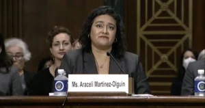 A woman is seated at a table with a nameplate that reads "Ms. Araceli Martínez-Olguín," suggesting she is participating in a formal hearing or meeting.