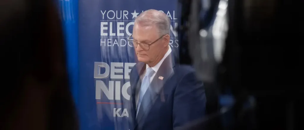 The image shows a man in a suit standing in front of a banner that reads "YOUR LOCAL ELECT HEADS DEF NICK KA."