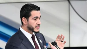 A man in a suit and tie is speaking into a microphone, gesturing with his hand.