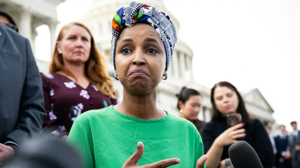A woman in a green shirt and colorful headscarf speaks to reporters in front of a historic building.