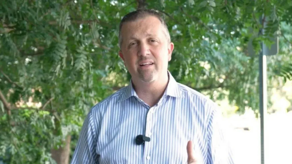 The image shows a man in a striped shirt standing outdoors with trees in the background, gesturing with his hand.