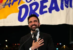 A man in a suit smiles while speaking into a microphone in front of a banner that reads "DREAM."