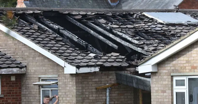 The photo shows a house with a damaged roof, likely after a fire, with visible structural damage and debris.