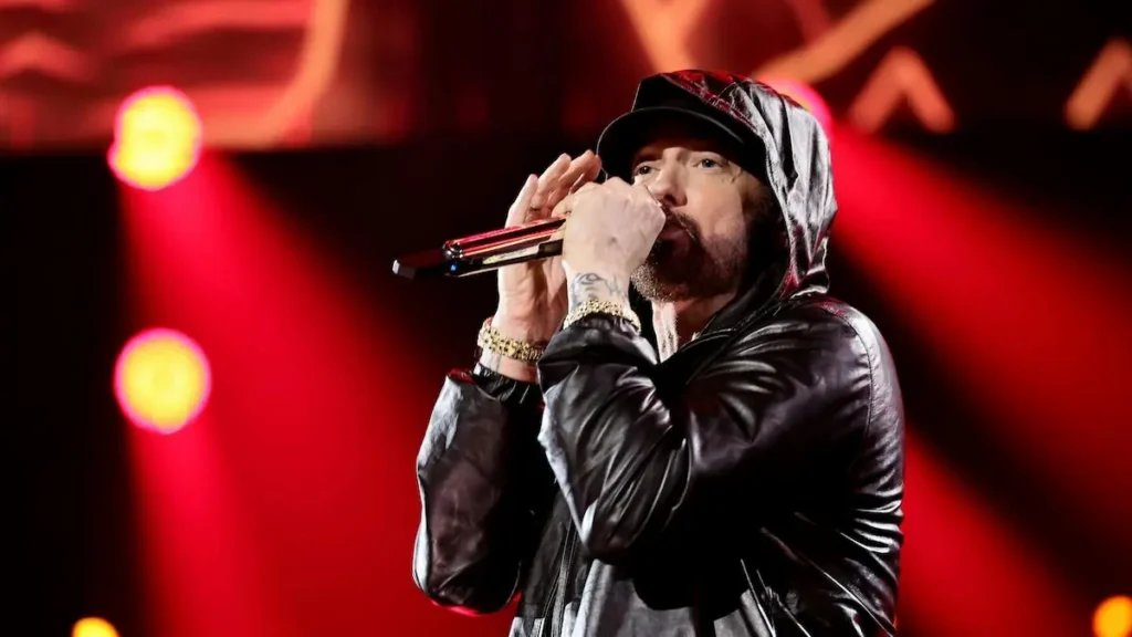 The image shows a performer on stage, wearing a black leather jacket and a cap, holding a microphone and singing into it.