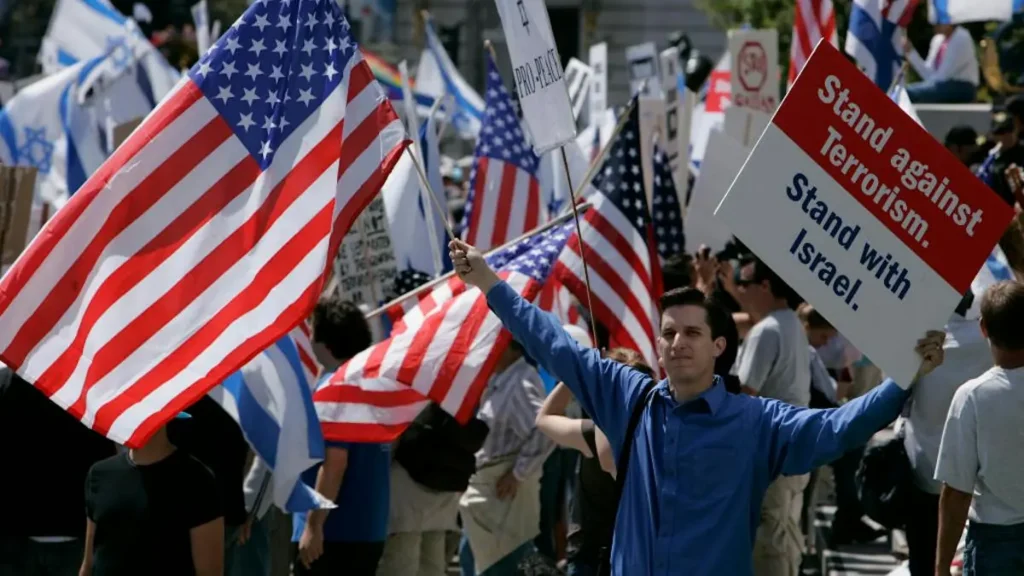 A man holds an American flag and a sign that reads "Stand against Terrorism. Stand with Israel" during a demonstration.