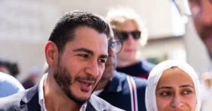 A man with a beard and a woman wearing a hijab are seen in a crowd, with other individuals in the background.