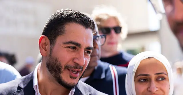 A man with a beard and a woman wearing a hijab are seen in a crowd, with other individuals in the background.