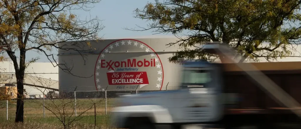 The image shows a large ExxonMobil sign celebrating "35 Years of Excellence" at the Joliet Refinery, with a blurred vehicle passing in the foreground.