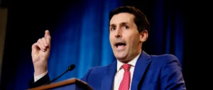 A man in a blue suit and red tie is speaking at a podium with a microphone, gesturing with his right hand.