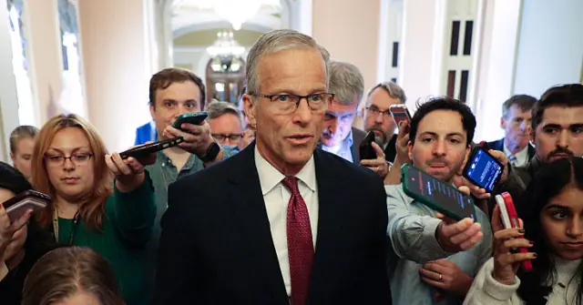 A man in a suit and tie is surrounded by reporters holding up their phones to record or take pictures.
