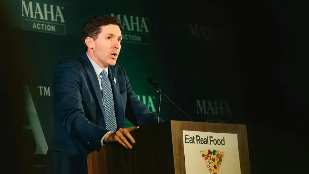 A man in a suit stands at a podium with a sign that reads "Eat Real Food," speaking at an event sponsored by MAHA Action.
