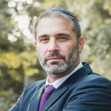 The image shows a man in a suit and tie, wearing a kippah, with a natural outdoor background.