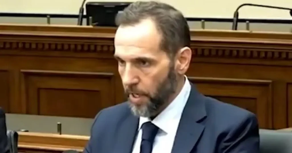 The image shows a man in a suit and tie, seated in what appears to be a formal setting, possibly a courtroom or a legislative chamber.