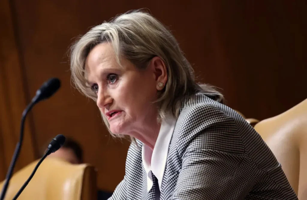The image shows a woman speaking at a formal event, likely a press conference or a public meeting, as indicated by the microphones in front of her.