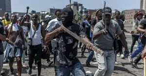 The image depicts a group of individuals engaged in a protest or demonstration, some holding sticks and wearing masks, suggesting a tense or confrontational situation.