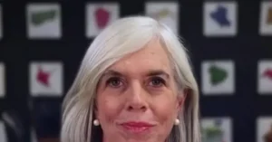 The image shows a person with long, white hair, wearing pearl earrings, in front of a backdrop featuring various symbols.