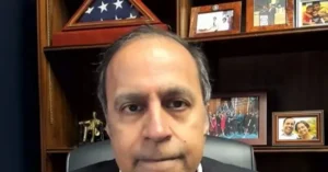 The image shows a man seated in an office with a backdrop featuring framed photos, a U.S. flag, and a group photo, suggesting a professional or political setting.