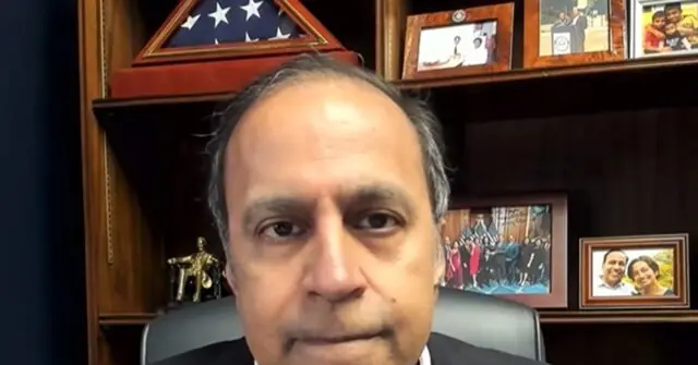 The image shows a man seated in an office with a backdrop featuring framed photos, a U.S. flag, and a group photo, suggesting a professional or political setting.