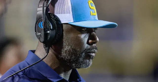 The image shows a person wearing a blue cap with a "S" logo and a headset, likely a coach or team official, observing a game or event.