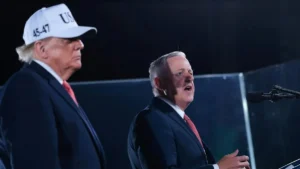 The image shows two individuals at a podium, one wearing a hat with "45-47" and the other speaking into a microphone.