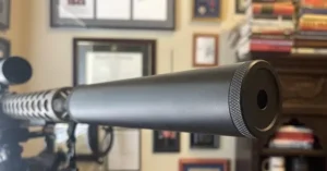 The image shows a close-up of a firearm's suppressor mounted on a rifle, with a background featuring framed certificates and books.