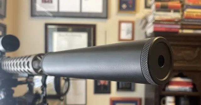 The image shows a close-up of a firearm's suppressor mounted on a rifle, with a background featuring framed certificates and books.