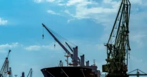 A large cargo ship is docked at a port, surrounded by industrial cranes under a partly cloudy sky.