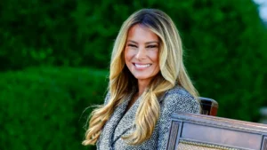 A woman with long, wavy blonde hair is smiling while seated outdoors, with a blurred green background.