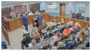 The image shows a public meeting or hearing taking place in a courtroom setting, with individuals seated in the audience and others standing or seated in the courtroom.
