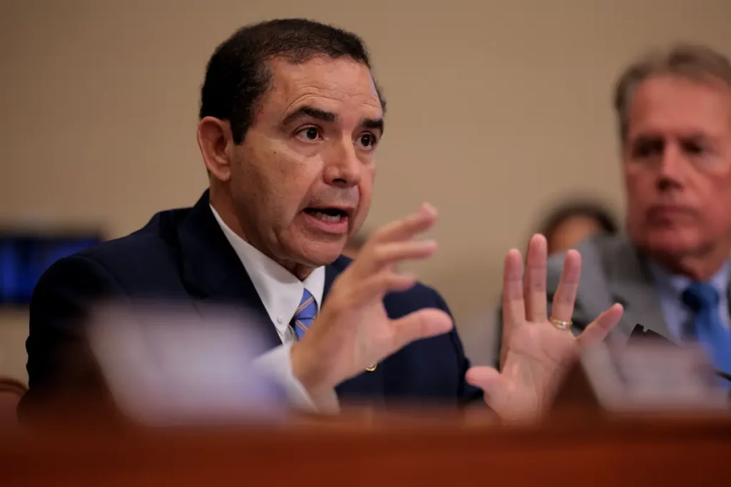 A man in a suit is speaking passionately with his hands raised during a formal meeting or hearing.