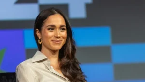 The image shows a person with long dark hair, wearing a light-colored shirt and gold earrings, seated in front of a colorful background.