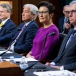 A group of individuals, including a woman in a purple top, are seated at a table with microphones, suggesting a formal meeting or hearing.