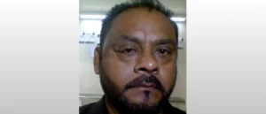 The image shows a man with a beard and mustache, wearing a dark shirt, in what appears to be an indoor setting with a light-colored wall and some papers or notices in the background.