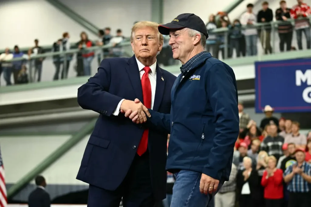 The image shows two individuals shaking hands at a public event, with a crowd of spectators in the background.