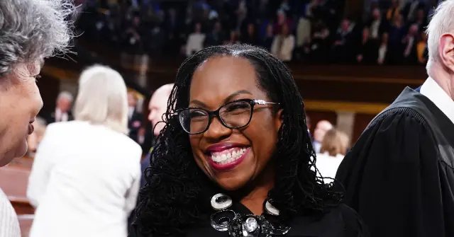 A woman smiles warmly at a gathering, surrounded by other attendees in a formal setting.