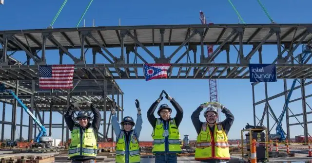 A group of construction workers celebrate at the Intel Foundry site, with flags and a clear blue sky in the background.