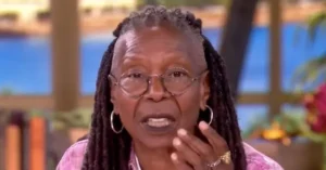 The image shows a person with long braided hair, wearing glasses and a pink top, speaking with their hand near their mouth.