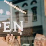 A person is holding up an "OPEN" sign in front of a store window, indicating the business is now open for business.