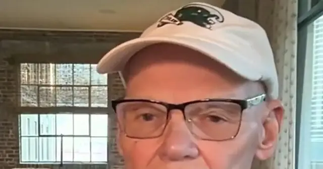The image shows a person wearing a white cap with a logo and glasses, standing indoors with a window and brick wall in the background.