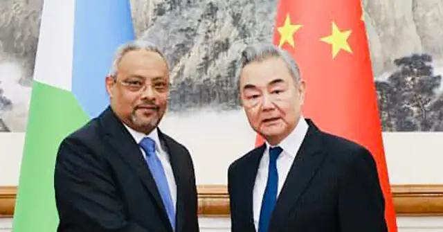 Two men in formal attire pose for a photo in front of flags, suggesting a diplomatic or official meeting.