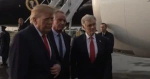 The image shows a group of men, including one in a suit with a red tie, walking near an aircraft with the U.S. presidential seal visible.