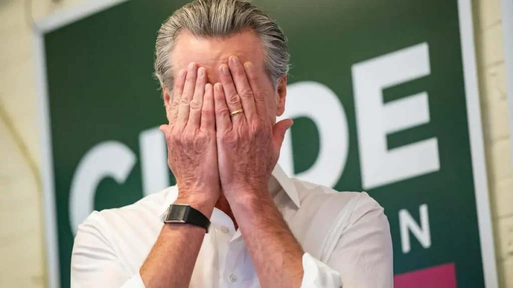 A man covers his face with his hands in front of a sign that reads "CUPDE."