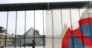 The image shows the Louvre Pyramid in Paris, viewed through a metal fence with a blurred flag in the foreground.