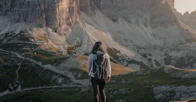 A hiker stands at the edge of a mountain, gazing at the rugged, snow-dusted peaks and a distant building.