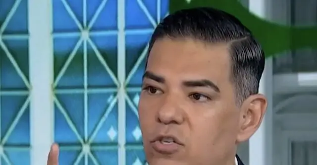 The image shows a man speaking in a news studio setting, with a geometric patterned background.