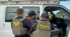 The image shows law enforcement officers from Immigration and Customs Enforcement (ICE) standing outside a vehicle.