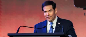 A man in a suit and tie is speaking at a podium with microphones, set against a red background.