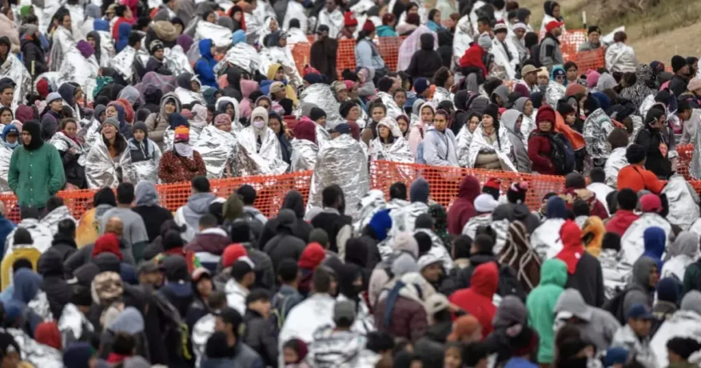 A large crowd of people, many wrapped in emergency blankets, are gathered behind orange fencing in what appears to be a humanitarian or relief situation.