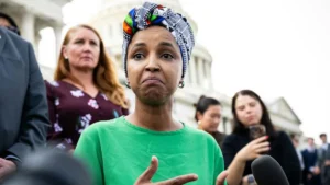 A woman in a green shirt and colorful headscarf speaks to reporters in front of a large building.