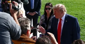The image shows a man in a suit and red tie speaking to a group of reporters and photographers in an outdoor setting.